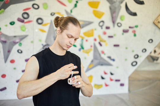 Young adult Caucasian man wrapping fingers with sports tape before bouldering session at climbing gym, preparing for grip support and injury prevention