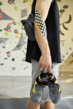 Vertical shot of young adult man holding climbing shoes beside taped forearm in bouldering gym, preparing for training