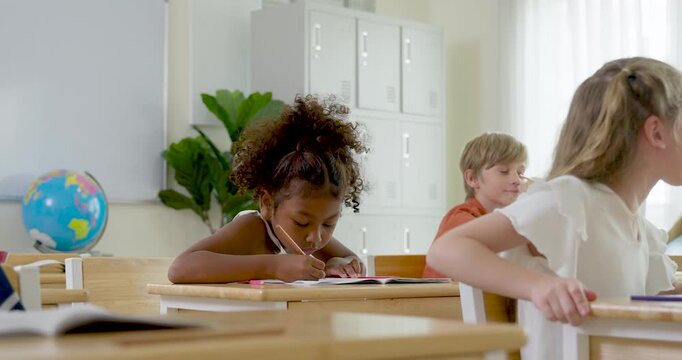 Multicultural children work at wooden desks with notebooks and school supply while chatting, listening and completing classwork during first day elementary classroom routine with teacher guidance