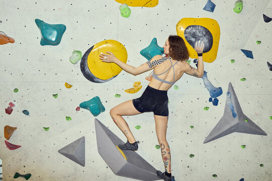 Young adult Caucasian woman climbing indoor bouldering wall, gripping holds and balancing on volumes, showing fitness challenge and active lifestyle