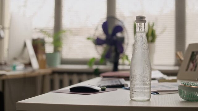 Medium shot of glass bottle of cold water placed on wooden desk in spacious office. Unrecognizable black woman working on computer in blurred background