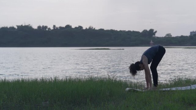A slow-motion of a South Asian woman in a standing forward bend (Uttanasana) doing yoga on a mat by a lake in tall grass.