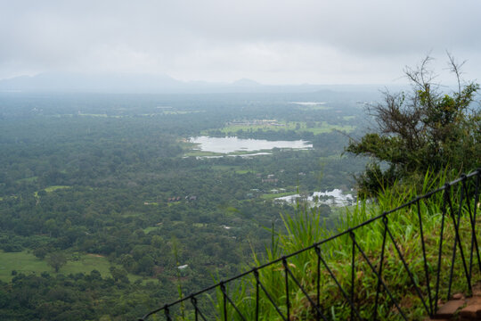 View of the lush green jungle landscape and a calm lake seen from a high vantage point with misty mountains under an overcast sky Sigiriya, Central Province, Sri Lanka.