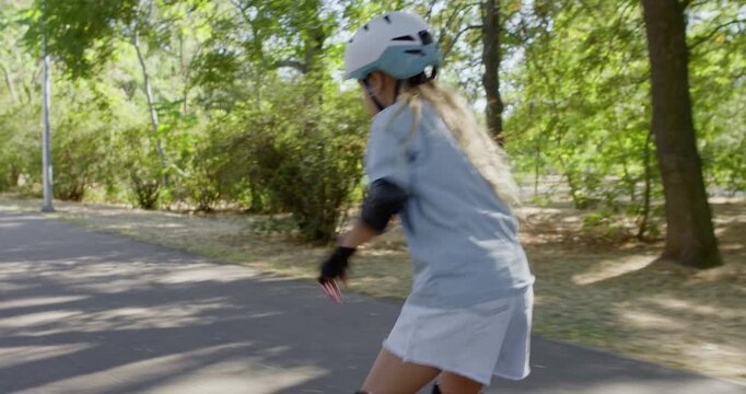 Girl skates along park path wearing helmet and protective pads. Child opens arms for balance keeping inline rollerblade movement controlled on paved path during ride
