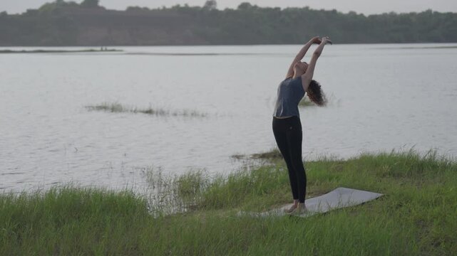 A slow-motion of a South Asian woman in a standing forward bend (Uttanasana) doing yoga on a mat by a lake in tall grass