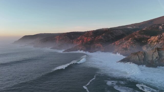 Drone flying above the Atlantic coastline near Guincho, Portugal, capturing long ocean waves rolling one after another toward the shore. The mountain ridge glows in golden sunset light as the drone sm
