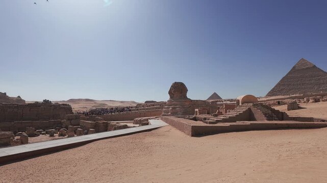 Wide angle handheld footage of the Great Sphinx surrounded by ancient stone structures with the pyramids of Giza rising in the background beneath a clear blue sky
