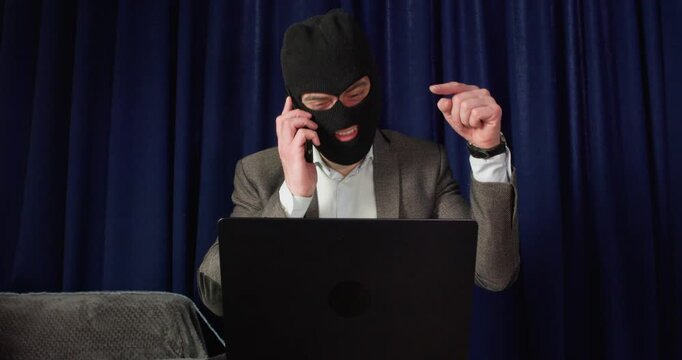 Masked male scammer talks on phone while working at laptop. Man points finger toward keyboard giving instructions during online extortion attempt from desk