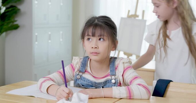 Young asian girl writes in notebook with pencil at wooden desk while classmate walks nearby, practicing assignment, attention, calm back to school study habit in bright primary classroom today.