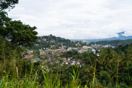 View of the urban landscape nestled among lush green hills and tropical trees under a cloudy sky in Kandy, Central Province, Sri Lanka.