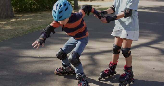 Girl helps younger brother stand up after rollerblading fall on park path. Sister grips hands steadying boy as skates slide across pavement during recovery
