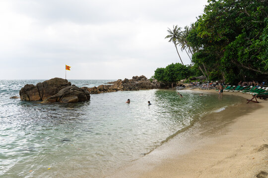 View of Secret Beach Mirissa with people swimming in clear water, rocky formations with a flag, and sun loungers under palm trees in Mirissa, Southern Province, Sri Lanka.