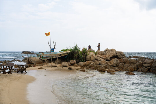 View of Secret Beach Mirissa featuring a traditional boat with a Sri Lankan flag on a rocky outcrop and people standing by the ocean in Mirissa, Southern Province, Sri Lanka.
