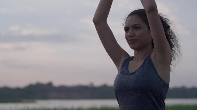 A Smiling South Asian woman with curly hair performing prayer hands pose (Anjali Mudra) during outdoor yoga by a lake at sunset