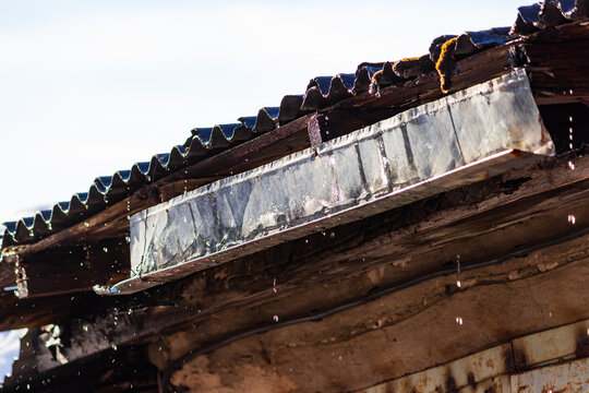 Clear water droplets dripping from an old weathered garage eaves edge, spring thaw melting concept with liquid splashes against a blurred rustic backdrop in bright daylight environment