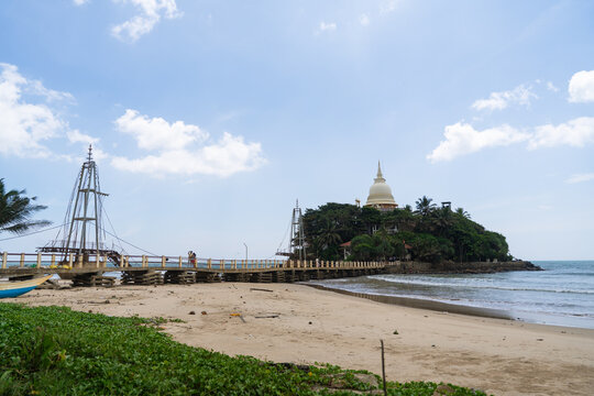 View of the Parevi Duwa Temple on a small island connected by a footbridge to the sandy beach under a blue sky Matara, Southern Province, Sri Lanka.