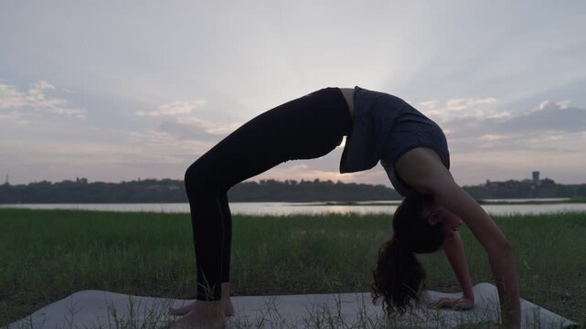 A slow-motion of a South Asian Woman in backbend doing yoga by a calm lake at twilight