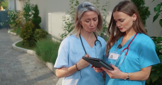 Two nurses discussing work and using digital tablet outside modern clinic. Professional healthcare teamwork and medical communication concept.
