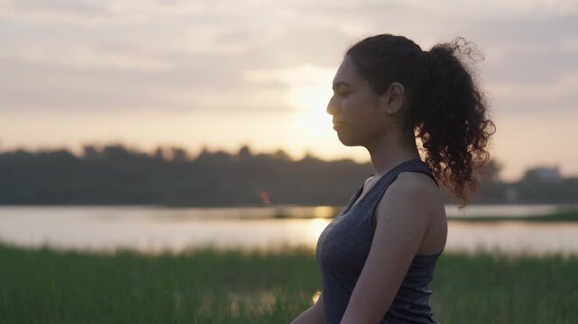 A wide-angle shot of a South Asian woman performing a graceful backbend on her yoga mat beside a quiet lake at sunrise