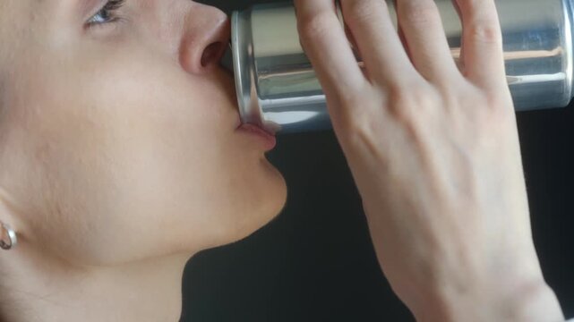 Side view close up young caucasian woman drinking refreshing beverage from silver aluminum can, savoring taste and quenching her thirst on solid black background. Woman enjoying refreshing soda drink