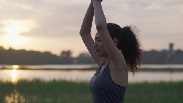 A South Asian woman practices yoga near a lake at sunrise, standing in a peaceful prayer position