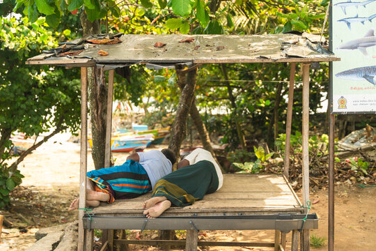 View of two men resting on a wooden platform under a makeshift shelter surrounded by lush tropical trees and a fishing sign in Galle, Southern Province, Sri Lanka.