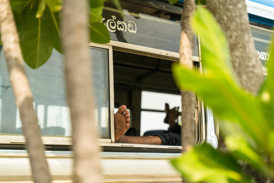 View of a passenger's bare feet resting on the window frame of a local bus framed by lush green tropical leaves in Galle, Southern Province, Sri Lanka.