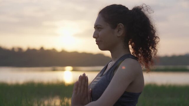 A side view of a South Asian woman seated in a meditative lotus pose on green grass at sunrise beside a calm reflective lake