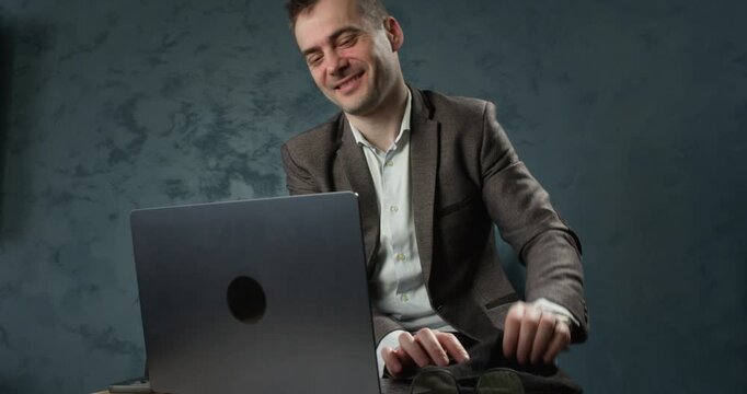 Scammer types on laptop and puts on mask after phone call during online fraud setup. Man hides face under balaclava while working on computer for deceptive scheme