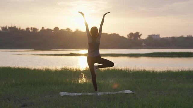 A woman practicing tree pose yoga during sunset on grassy riverside with peaceful golden light