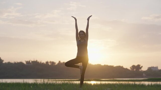 A South Asian woman holding a balanced tree pose beside a calm lakeside, the rising sun casting a warm golden glow
