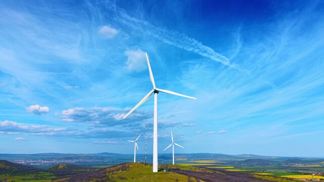 White wind turbines on grassy hill with blue sky background. Wide angle view of alternative energy wind turbines on a green hill against a vibrant blue sky with cirrus clouds.