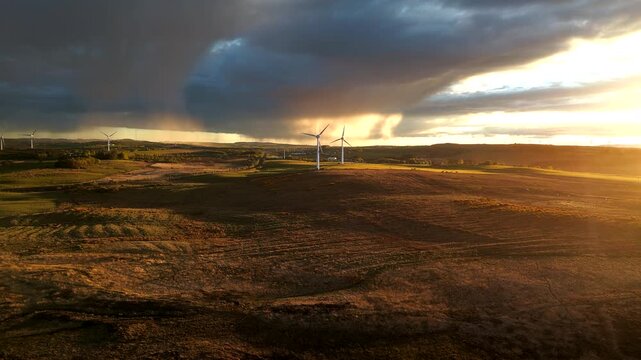 Wide drone footage of wind turbines in open countryside with fields, dark clouds, and warm sunset light in Scotland.