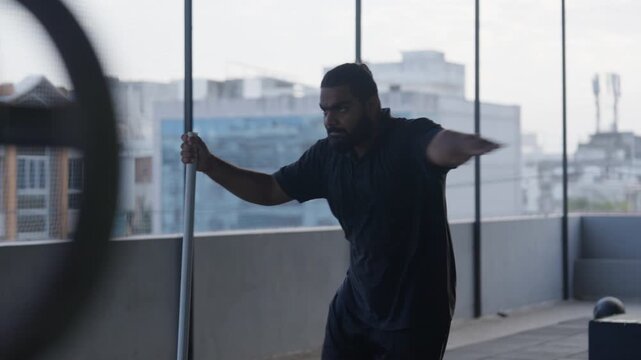 A focused Indian bearded athlete in dark sportswear performs dynamic leg swings as pre-workout warm up in a floor-to-ceiling windowed modern gym overlooking a city skyline. Smooth cinematic right-to-l