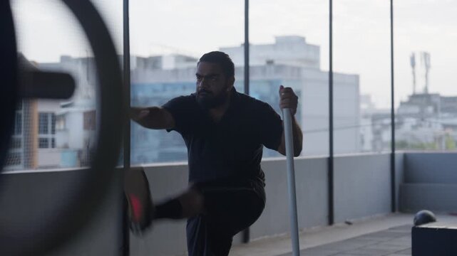 A focused Indian bearded athlete in dark sportswear performs dynamic leg swings as pre-workout warm up in a floor-to-ceiling windowed modern gym overlooking a city skyline. Smooth cinematic right-to-l