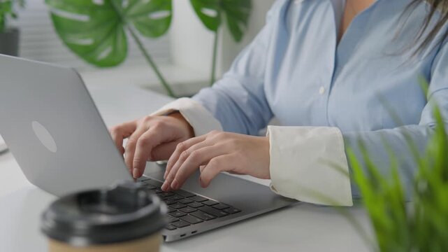 Closeup of womans hands typing on laptop in bright workspace with coffee and greenery, symbolizing remote work trends. Modern office environment highlights technology and flexible work lifestyle