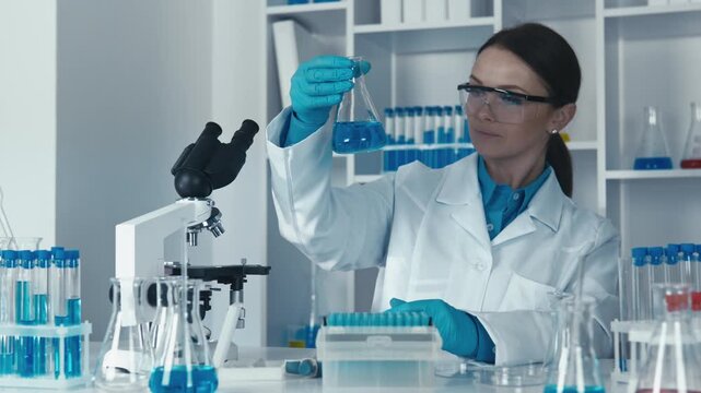 A woman researcher conducts scientific analysis in a high tech lab, examining a blue solution in a glass flask. Laboratory equipment and safety gear emphasize innovation and precision