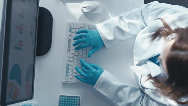 A researcher in a white lab coat and blue gloves types on a keyboard at a laboratory workstation, surrounded by scientific charts and equipment. The image captures innovation and technology in science