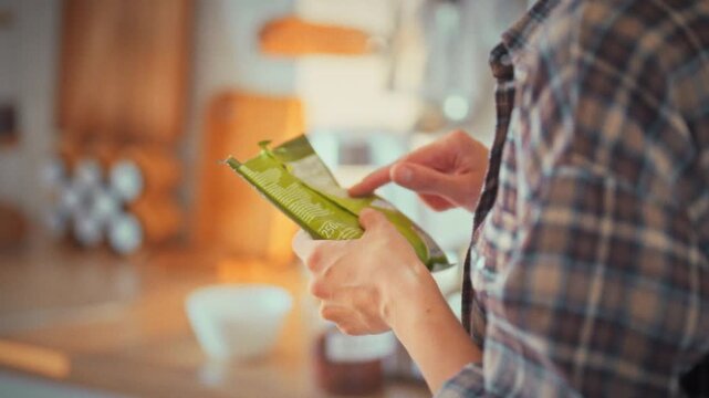 In a bright home kitchen at midday, a girl inspects the soft plastic packaging of a food product, scrutinizing its composition to ensure proper nutrition and make a conscious, healthy food choice