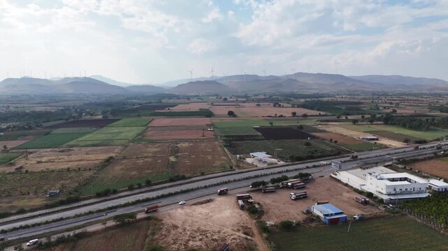 Drone shot capturing the highway corridor near Chitradurga with expansive agricultural fields, industrial structures, and rolling mountain ranges lined with windmills beneath dramatic skies.