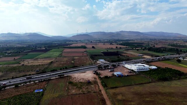 Cinematic aerial view of the Mumbai Highway passing through the rural landscapes of Chitradurga, surrounded by patchwork farmlands and distant wind turbine-covered hills.
