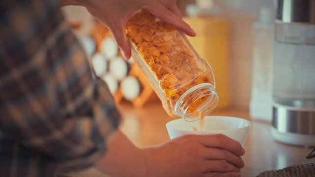 Woman gently pours cornflakes into bowl in her bright kitchen, assembling light breakfast. The close-up highlights her hands and the crisp cereal, emphasizing proper nutrition and a healthy lifestyle