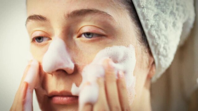 In a bright bathroom, a girl massages white cleansing foam over her facial skin, gently lifting makeup and impurities. This quick skincare step refreshes her complexion and readies her for the day