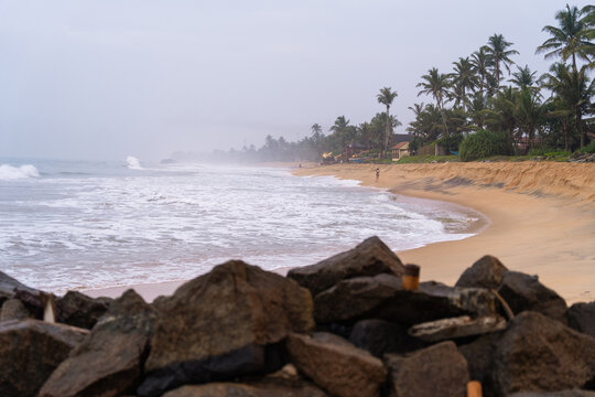 View of a sandy tropical beach with crashing waves, palm trees lining the shore, and dark rocks in the foreground under an overcast sky in Galle, Southern Province, Sri Lanka.