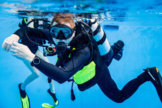 Close-up underwater view of a male scuba diver in a pool looking forward with another diver partially visible in the background