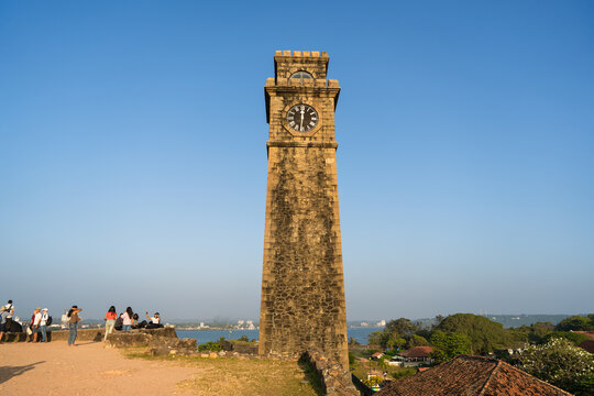 View of the historic Galle Fort Clock Tower with tourists standing on the ramparts under a clear blue sky in Galle, Southern Province, Sri Lanka.