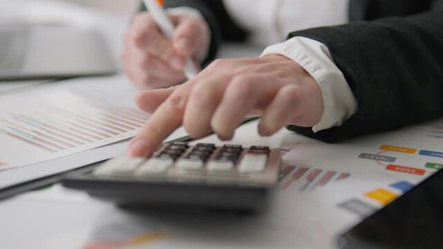 Focused person reviews financial graphs and calculates data for business analysis and strategy. Office workspace features paperwork, pen, and calculator for accurate financial management
