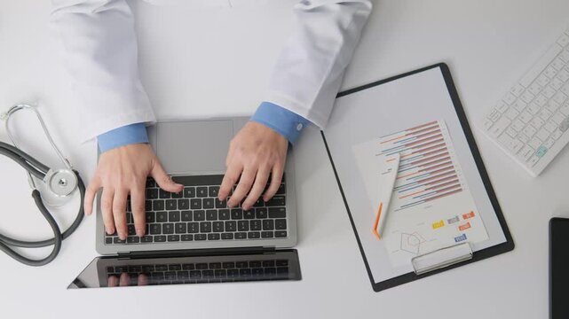 A physician types on a laptop surrounded by medical documents, a stethoscope, and analytical charts. Digital healthcare solutions and data driven decision making are highlighted in this workspace