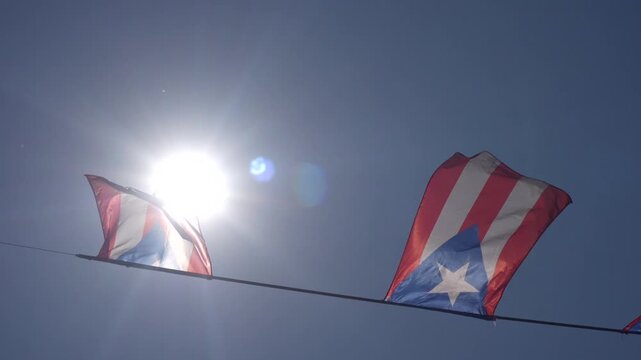 A string of Puerto Rican flags waving in the wind against a clear blue sky with sun rays.
