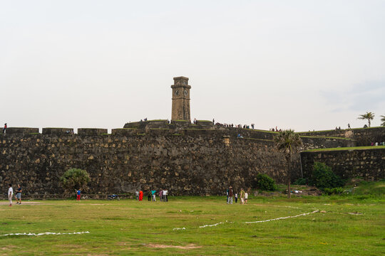 View of the historic Galle Fort Clock Tower rising above massive stone ramparts with people walking on the grass and walls Galle, Southern Province, Sri Lanka.